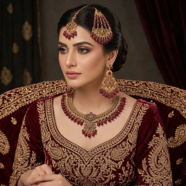 Gold-Tone Ruby Red Bead Filigree Choker & Chandbali Bridal Jewelry Set worn on Pakistani female bridal model in deep maroon velvet lehenga seated on ornate bridal chair with burgundy curtain backdrop