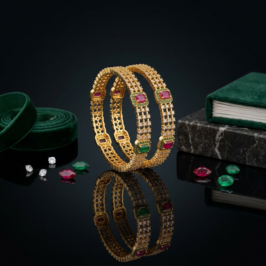 22K Gold-Tone Ruby Red CZ Cushion Bezel & Emerald Green Pavé Border Honeycomb Filigree Openwork & Triple-Row White CZ Wide Bangle Set of 2 on black mirror surface with loose ruby and emerald CZ stones, green velvet ribbon and dark marble book prop backdrop