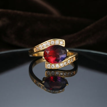 Gold Ruby Red Oval CZ Bypass Crossover Cocktail Ring on dark grey mirror surface with dark brown branches backdrop