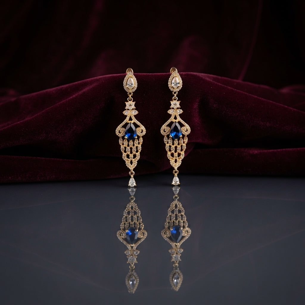 Gold Sapphire Blue CZ Filigree Chandelier Drop Earrings — 18-22K gold-tone Art Deco bridal chandelier earrings with pear CZ halo stud snowflake CZ connector elongated open gold filigree lattice body with sapphire blue pear CZ center focal and single pear CZ halo drop displayed on dark grey mirror surface with burgundy velvet backdrop