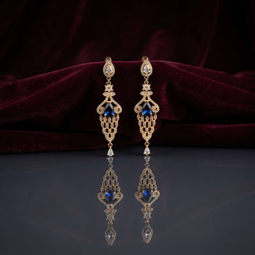 Gold Sapphire Blue CZ Filigree Chandelier Drop Earrings — 18-22K gold-tone Art Deco bridal chandelier earrings with pear CZ halo stud snowflake CZ connector elongated open gold filigree lattice body with sapphire blue pear CZ center focal and single pear CZ halo drop displayed on dark grey mirror surface with burgundy velvet backdrop