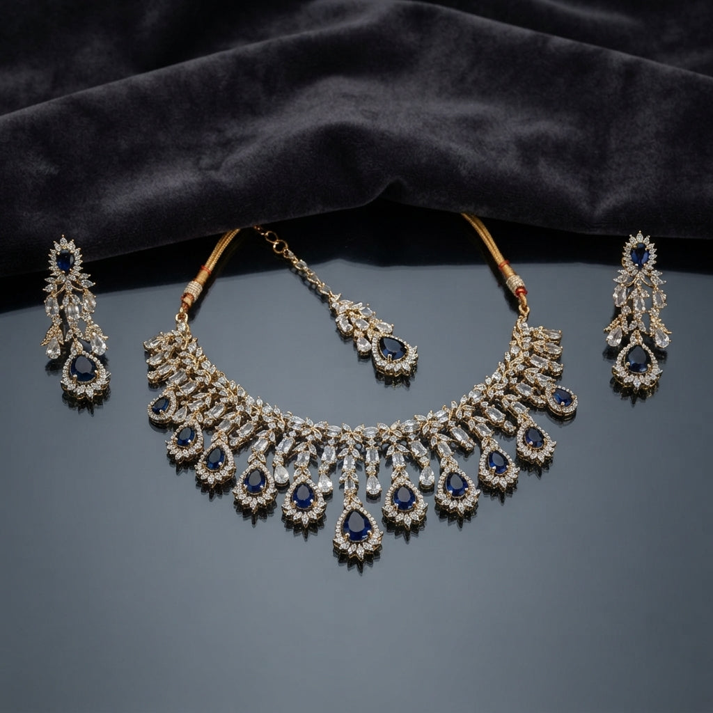 Gold Sapphire Blue CZ Leaf Cascade Necklace Maang Tikka and Linear Drop Earrings Bridal Set — 22K gold-tone 3-piece bridal set with graduating sapphire blue pear CZ halo cascade fringe necklace with marquise white CZ leaf clusters and baguette fans matching maang tikka with sapphire blue pear CZ halo drop and 4-tier linear cascade drop earrings displayed on dark grey mirror surface with charcoal satin backdrop