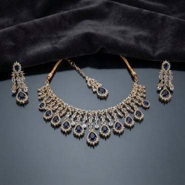 Gold Sapphire Blue CZ Leaf Cascade Necklace Maang Tikka and Linear Drop Earrings Bridal Set — 22K gold-tone 3-piece bridal set with graduating sapphire blue pear CZ halo cascade fringe necklace with marquise white CZ leaf clusters and baguette fans matching maang tikka with sapphire blue pear CZ halo drop and 4-tier linear cascade drop earrings displayed on dark grey mirror surface with charcoal satin backdrop