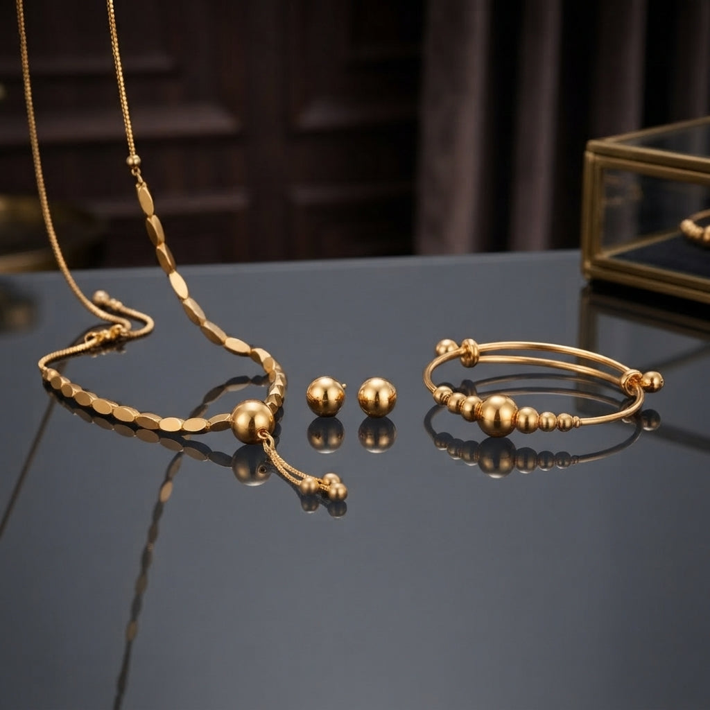 Gold Smooth Ball Bead Tassel Necklace Bracelet Earrings Set 3 Piece in 22K gold-tone plating on dark grey blue mirror surface with dark interior and gold glass jewellery box backdrop showing flat oval link chain necklace with large smooth gold ball drop pendant double-wire expandable bangle with smooth graduated gold ball beads and large smooth round gold dome ball stud earrings with mirror reflections below