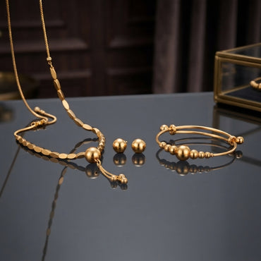 Gold Smooth Ball Bead Tassel Necklace Bracelet Earrings Set 3 Piece in 22K gold-tone plating on dark grey blue mirror surface with dark interior and gold glass jewellery box backdrop showing flat oval link chain necklace with large smooth gold ball drop pendant double-wire expandable bangle with smooth graduated gold ball beads and large smooth round gold dome ball stud earrings with mirror reflections below