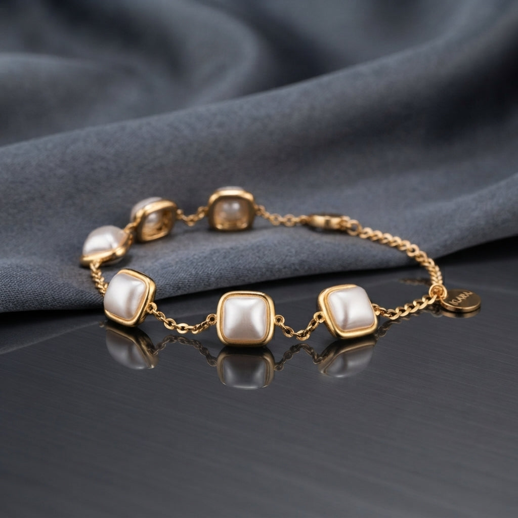 Gold Square Cushion Faux Pearl Bezel Station Bracelet flat lay on dark grey mirror surface with grey silk backdrop showing six square cushion faux pearl stations in polished gold-tone bezel settings linked on gold cable chain with lobster clasp and extension with mirror reflection below