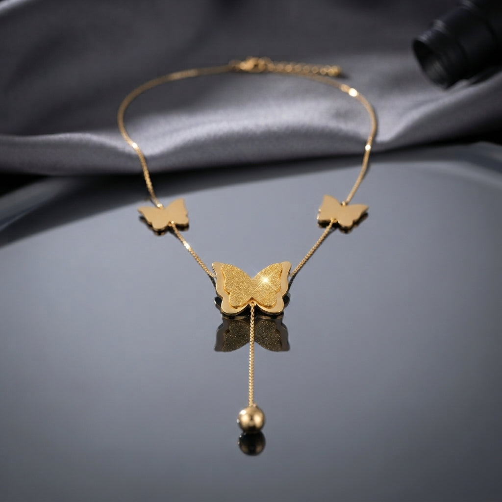 Gold Textured Butterfly Multi-Charm Station Tassel Ball Drop Necklace flat lay on grey mirror surface with grey silk backdrop showing two small smooth gold butterfly station charms flanking large textured filigree engraved center butterfly pendant with single tassel drop chain and polished gold ball charm with mirror reflection below