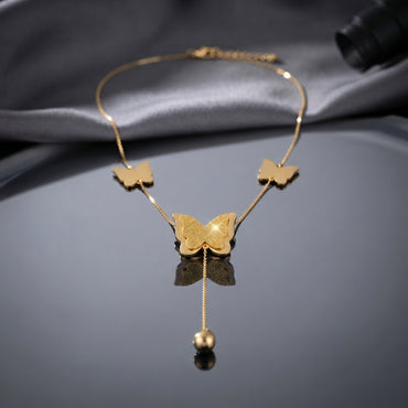 Gold Textured Butterfly Multi-Charm Station Tassel Ball Drop Necklace flat lay on grey mirror surface with grey silk backdrop showing two small smooth gold butterfly station charms flanking large textured filigree engraved center butterfly pendant with single tassel drop chain and polished gold ball charm with mirror reflection below