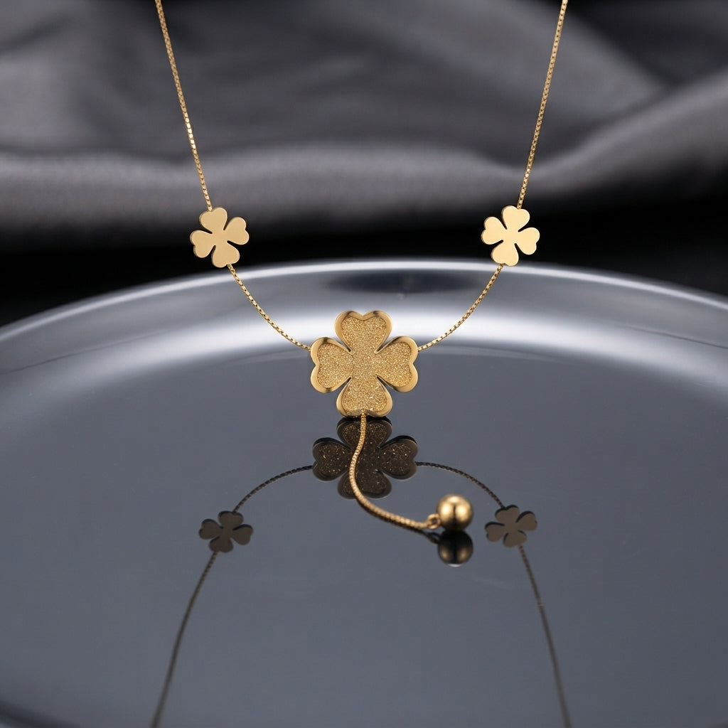 Gold Textured Four-Leaf Clover Multi-Charm Station Necklace flat lay on silver chrome circular surface with dark grey silk backdrop showing three smooth gold clover station charms on box chain with large textured glitter center clover pendant and gold ball drop closure with mirror reflection below