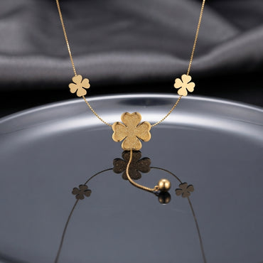 Gold Textured Four-Leaf Clover Multi-Charm Station Necklace flat lay on silver chrome circular surface with dark grey silk backdrop showing three smooth gold clover station charms on box chain with large textured glitter center clover pendant and gold ball drop closure with mirror reflection below