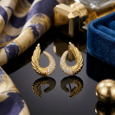 22K Gold-Tone Angel Wing Feather Crescent Pavé White CZ Stud Earrings pair on dark reflective surface with indigo blue floral scarf and gold cube perfume bottle backdrop