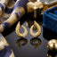 22K Gold-Tone Angel Wing Feather Crescent Pavé White CZ Stud Earrings pair on dark reflective surface with indigo blue floral scarf and gold cube perfume bottle backdrop