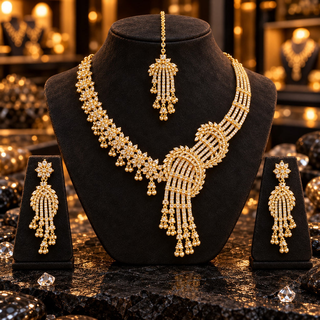 Gold-tone asymmetric necklace with clear floral clusters, parallel clear-accent strands, curved leaf-style frames and gold-tone bead tassels, beside matching long earrings and maang tikka on dark stands.