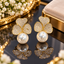 Gold-tone earrings with three clear-accent heart-shaped petals forming a clover motif and round ivory-white pearl-look details beneath.