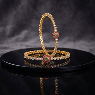 Gold-Tone CZ Tennis Slim Bangle Pair with Multi-Color Teardrop CZ Focal Clasp displayed upright and flat on black lacquer round stand with dark grey marble backdrop showing two matching slim gold bangles with continuous single row round white CZ prong-set stones and heart teardrop focal clasp with emerald-green CZ center ruby-red CZ halo and white CZ outer border with mirror reflection below