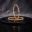 Gold-Tone CZ Tennis Slim Bangle Pair with Multi-Color Teardrop CZ Focal Clasp displayed upright and flat on black lacquer round stand with dark grey marble backdrop showing two matching slim gold bangles with continuous single row round white CZ prong-set stones and heart teardrop focal clasp with emerald-green CZ center ruby-red CZ halo and white CZ outer border with mirror reflection below