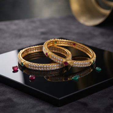 Gold-Tone Double-Row CZ Pave Multi-Color Oval CZ Accent Wide Bangle Pair stacked on black mirror tray with dark marble backdrop and scattered ruby and emerald CZ stones showing two matching wide gold bangles with two continuous rows of round white CZ pave stones gold sunburst fan engraved dividers and large oval ruby-red and emerald-green CZ accent stones in gold bezel settings with mirror reflection below