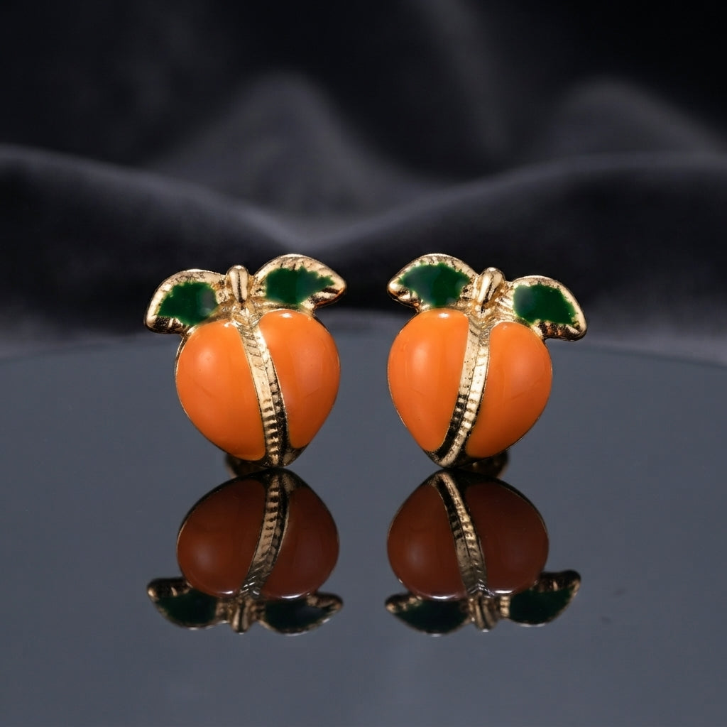 Gold-Tone Enamel Fruit Peach Stud Earrings in orange enamel pair displayed on dark mirror surface with dark silk drape backdrop showing round orange enamel peach form with gold-tone ribbed engraved seam divider and two green enamel leaves at top with mirror reflection below