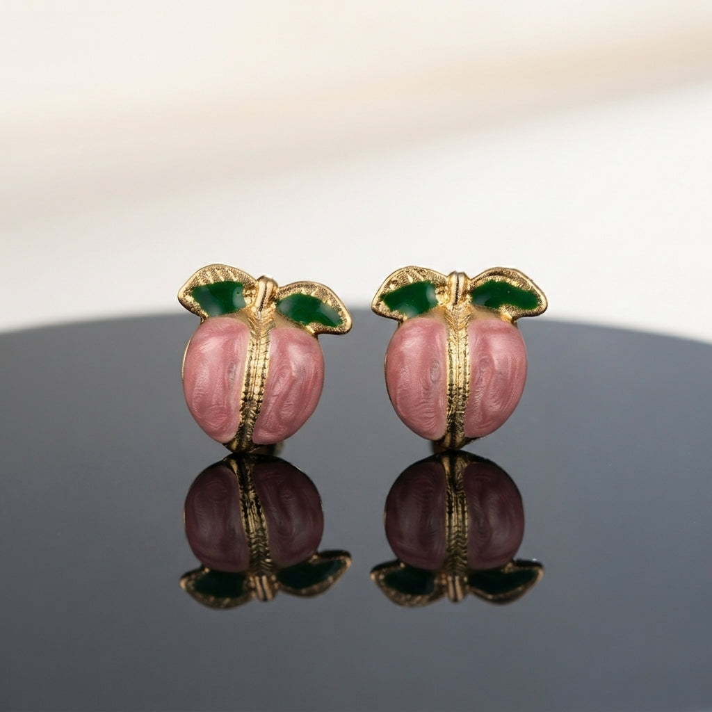 Gold-Tone Enamel Fruit Peach Stud Earrings in pink enamel pair displayed on dark grey mirror surface with white fabric backdrop showing round pink enamel peach form with gold-tone ribbed engraved seam and two green enamel leaves with mirror reflection below