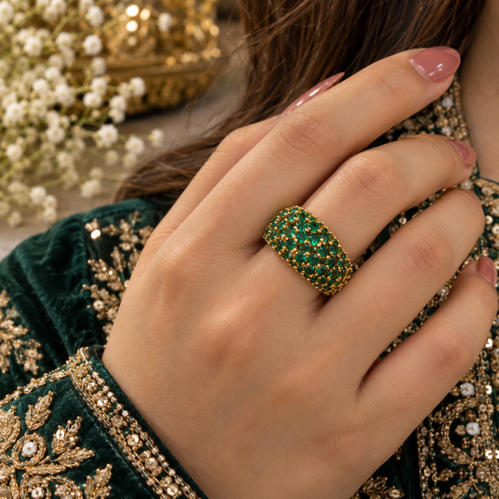 Model wearing a gold-tone wide dome ring with closely arranged green round accents and bead-like setting details.
