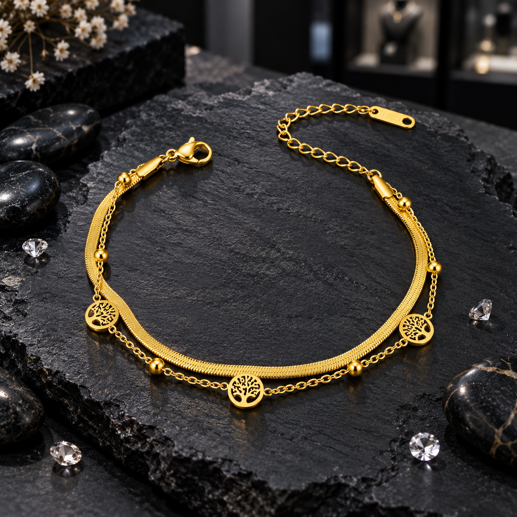Gold-tone layered bracelet with a flat textured chain, fine link chain, three round openwork tree charms, small beads, lobster-style clasp and extension chain on dark slate.
