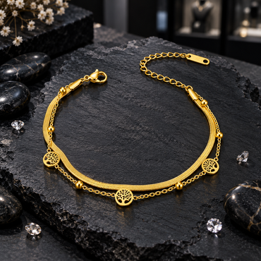 Gold-tone layered bracelet with a flat textured chain, fine link chain, three round openwork tree charms, small beads, lobster-style clasp and extension chain on dark slate.