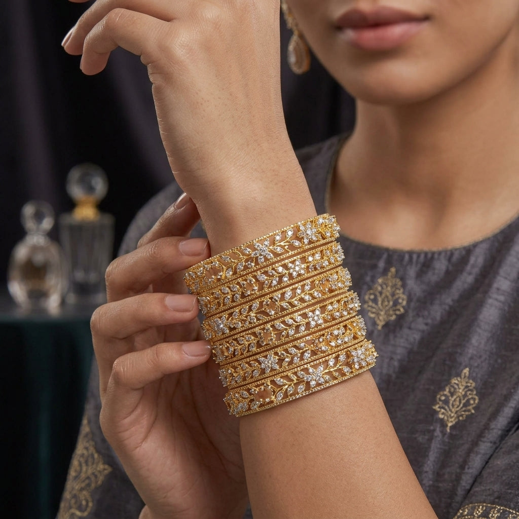 Gold-Tone Marquise and Round CZ Floral Leaf Open-Work Wide Bangle Set of 4 worn stacked on model wrist in grey embroidered outfit showing four matching wide gold bangles with intricate open-work filigree leaf vine lattice marquise CZ leaf-vein settings round CZ pave scatter and champagne pink CZ solitaire accent with crystal perfume bottles in background