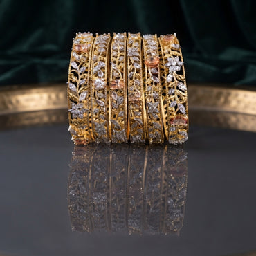 Gold-Tone Marquise and Round CZ Floral Leaf Open-Work Wide Bangle Set of 4 standing upright on dark grey mirror tray with emerald green velvet backdrop showing four matching wide gold bangles with open-work filigree leaf vine lattice marquise CZ settings round CZ pave and large oval champagne pink CZ solitaire accents with mirror reflection below