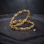 Gold-Tone Multi-Color CZ Floral Infinity Twist Slim Bangle Pair on dark slate stone surface with scattered ruby emerald and diamond CZ stones showing two matching slim gold bangles with continuous infinity twist gold links alternating with ruby-red and emerald-green CZ floral cluster rosettes with white CZ petals with mirror reflection below