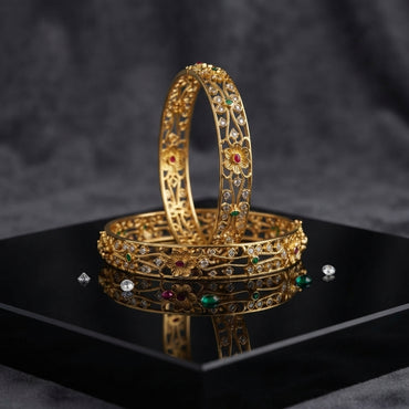 Gold-Tone Multi-Color CZ Scattered Floral Open-Work Filigree Wide Bangle Pair displayed upright and flat on black mirror tray with dark grey marble backdrop showing two matching wide gold bangles with intricate open-work filigree lattice vine branch motifs scattered round white CZ stones large 5-petal ruby-red CZ center daisy flowers and teardrop emerald-green CZ accents with mirror reflection below