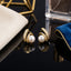 Gold-Tone Pearl Center Baguette & Round Pavé CZ Swirl Crescent Stud Earrings pair on dark reflective surface with gold geometric star ornament and crystal perfume bottle backdrop