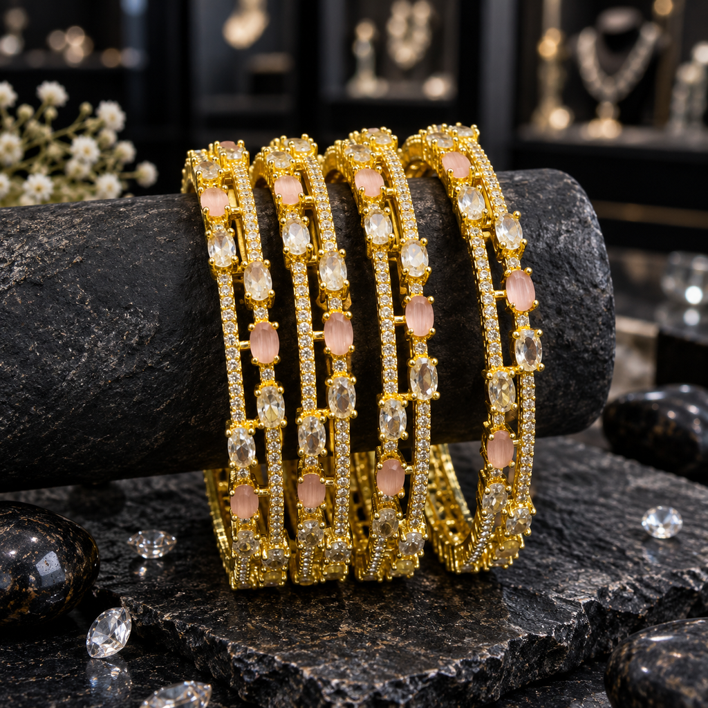 Four gold-tone AD bangles on a black display roll, with alternating pale pink and clear oval accents, slim clear accent rows and connecting bars.