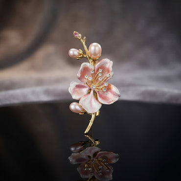 Gold-Tone Pink Enamel Cherry Blossom Faux Pearl CZ Stem Brooch Pin standing upright on dark black mirror surface with blurred dark wood and mist backdrop showing gold twig branch with large open 5-petal blush to deep pink gradient enamel cherry blossom flower with CZ wire stamen accents four cream faux pearl buds and small pink CZ bud tip with mirror reflection below