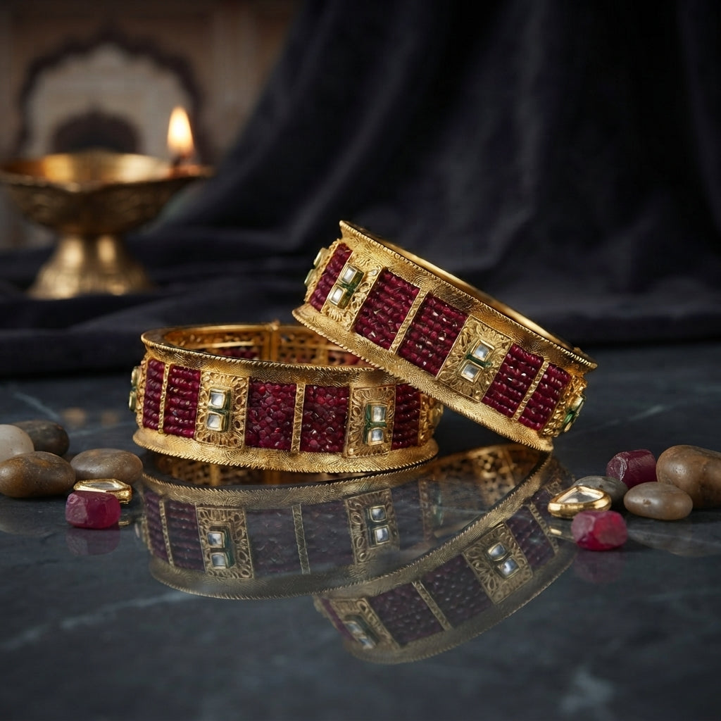 Pair of wide gold-tone bangles with deep red panels, square clear accents and ornate borders on a dark reflective surface.