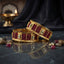 Pair of wide gold-tone bangles with deep red panels, square clear accents and ornate borders on a dark reflective surface.