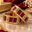 Two wide gold-tone bangles showing red panels, green-edged clear accents, textured borders and decorative openwork on marble.