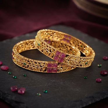 Gold-Tone Ruby Square Emerald Accent Floral Filigree Wide Bangle Pair stacked on dark slate stone surface with burgundy velvet backdrop and scattered ruby and emerald gemstones showing two matching wide gold bangles with intricate floral filigree open-work lattice body 4 square ruby-red CZ panel in 2x2 grid and small round emerald-green CZ flower center accents throughout