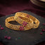 Gold-Tone Ruby Square Emerald Accent Floral Filigree Wide Bangle Pair stacked on dark slate stone surface with burgundy velvet backdrop and scattered ruby and emerald gemstones showing two matching wide gold bangles with intricate floral filigree open-work lattice body 4 square ruby-red CZ panel in 2x2 grid and small round emerald-green CZ flower center accents throughout
