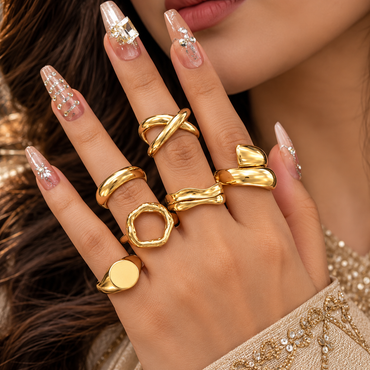 Six gold-tone sculptural rings worn across a model's fingers, showing crossover, domed, open-centre, double-wave, wrap-style and round signet-style designs.