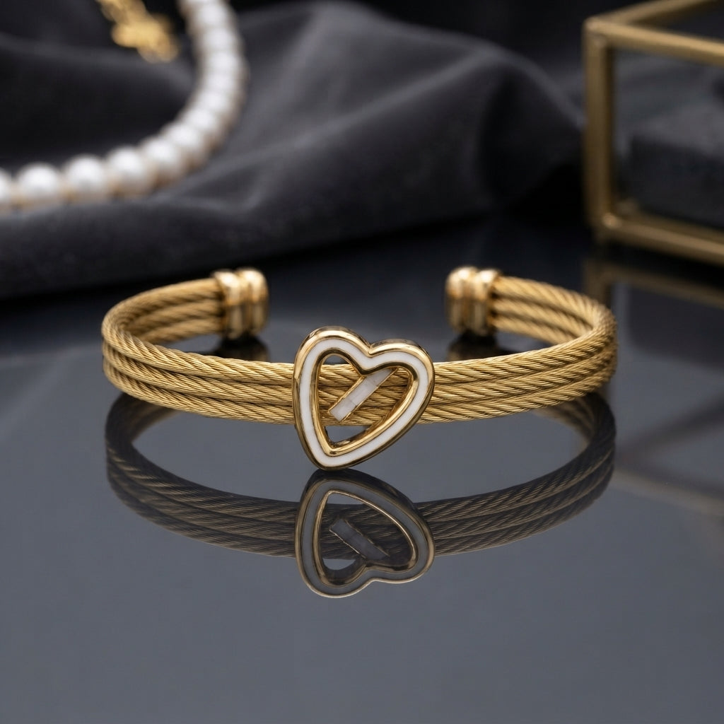 Gold Triple Cable Twist Heart Enamel Cuff Bangle in 18K gold-tone plated stainless steel on dark grey mirror surface with dark fabric and pearl necklace backdrop showing open-ended cuff with three twisted cable rope strands gathered by gold knot barrel connectors at each end and open heart focal with white enamel outline at centre with cable strands passing through heart and mirror reflection below
