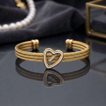 Gold Triple Cable Twist Heart Enamel Cuff Bangle in 18K gold-tone plated stainless steel on dark grey mirror surface with dark fabric and pearl necklace backdrop showing open-ended cuff with three twisted cable rope strands gathered by gold knot barrel connectors at each end and open heart focal with white enamel outline at centre with cable strands passing through heart and mirror reflection below