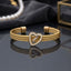 Gold Triple Cable Twist Heart Enamel Cuff Bangle in 18K gold-tone plated stainless steel on dark grey mirror surface with dark fabric and pearl necklace backdrop showing open-ended cuff with three twisted cable rope strands gathered by gold knot barrel connectors at each end and open heart focal with white enamel outline at centre with cable strands passing through heart and mirror reflection below