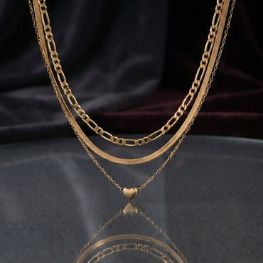 Gold Triple Layer Figaro Herringbone and Heart Pendant Chain Necklace — 18K gold-tone unified triple layered necklace with figaro chain, flat herringbone snake chain and delicate cable chain with 3D puffy gold heart pendant displayed as flat lay on dark satin surface with mirror reflection