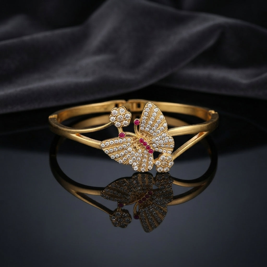 Gold White CZ and Ruby Red Butterfly Pave Hinged Cuff Bangle product flat lay standing upright on dark black mirror surface with dark satin velvet backdrop showing 22K warm yellow gold-tone hinged cuff bangle with large 3D sculptural butterfly featuring dense micro-pave white CZ wings engraved vein texture ruby red CZ body pave CZ flower antennae tips and side flower accents with sharp mirror reflection below