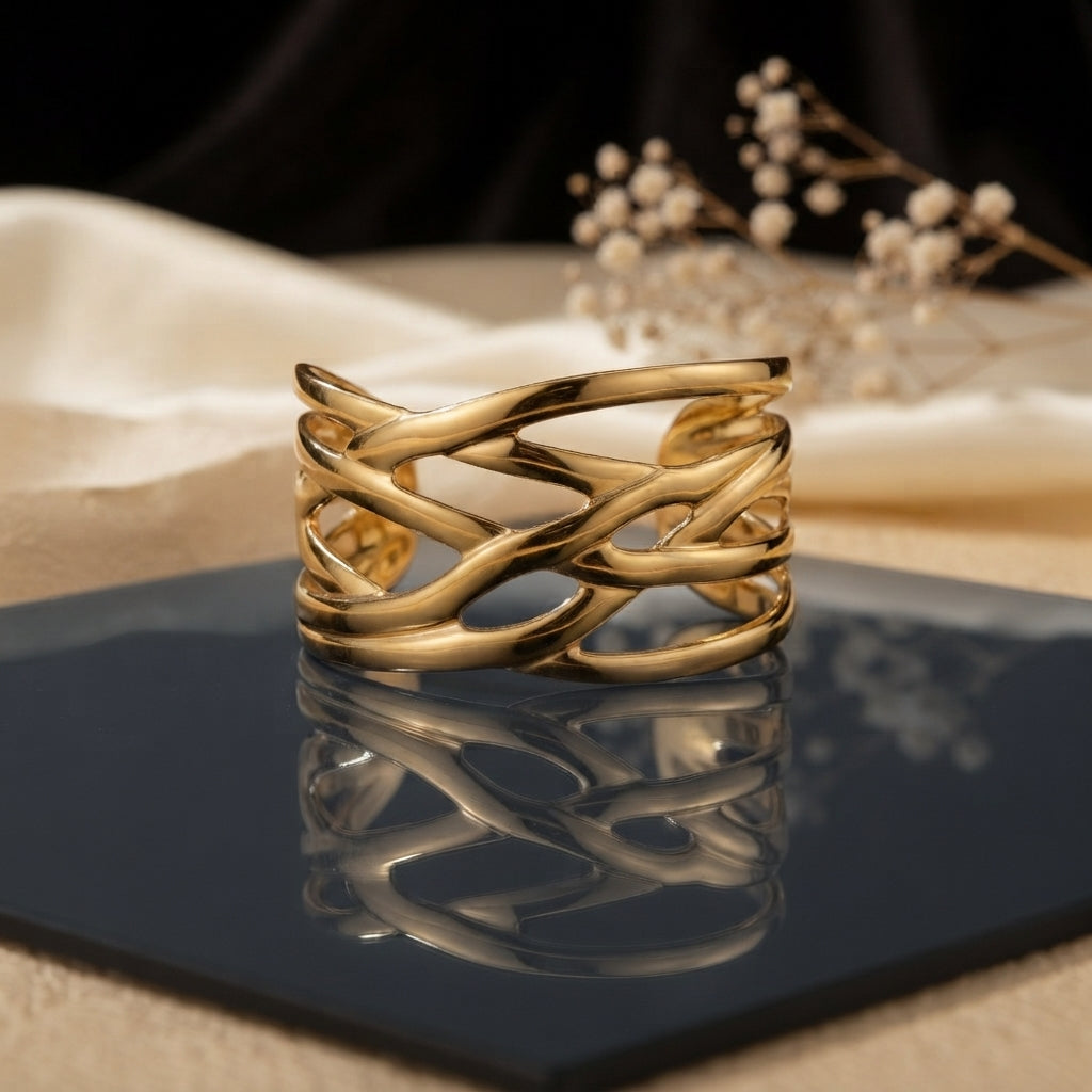 Gold Wide Braided Wire Weave Open Cuff Bangle in 18K gold-tone PVD mirror polish stainless steel on dark navy blue ceramic tile with cream silk fabric and white dried flowers backdrop showing wide open cuff with 4-5 slim smooth wire strands in continuous diagonal braid criss-cross weave pattern with open diamond eye-shaped negative spaces and mirror reflection below