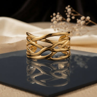Gold Wide Braided Wire Weave Open Cuff Bangle in 18K gold-tone PVD mirror polish stainless steel on dark navy blue ceramic tile with cream silk fabric and white dried flowers backdrop showing wide open cuff with 4-5 slim smooth wire strands in continuous diagonal braid criss-cross weave pattern with open diamond eye-shaped negative spaces and mirror reflection below