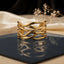 Gold Wide Braided Wire Weave Open Cuff Bangle in 18K gold-tone PVD mirror polish stainless steel on dark navy blue ceramic tile with cream silk fabric and white dried flowers backdrop showing wide open cuff with 4-5 slim smooth wire strands in continuous diagonal braid criss-cross weave pattern with open diamond eye-shaped negative spaces and mirror reflection below