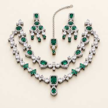 Emerald-green AD bridal jewelry set with layered silver-tone necklaces, matching earrings, and maang tikka on an ivory studio background