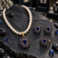 Cream pearl-look beaded necklace with dark-blue oval pendant, matching earrings and ring, surrounded by blue decorative accents on dark slate.