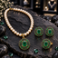 Cream pearl-look beaded necklace with green oval pendant, matching earrings and ring, surrounded by green decorative accents on dark slate.